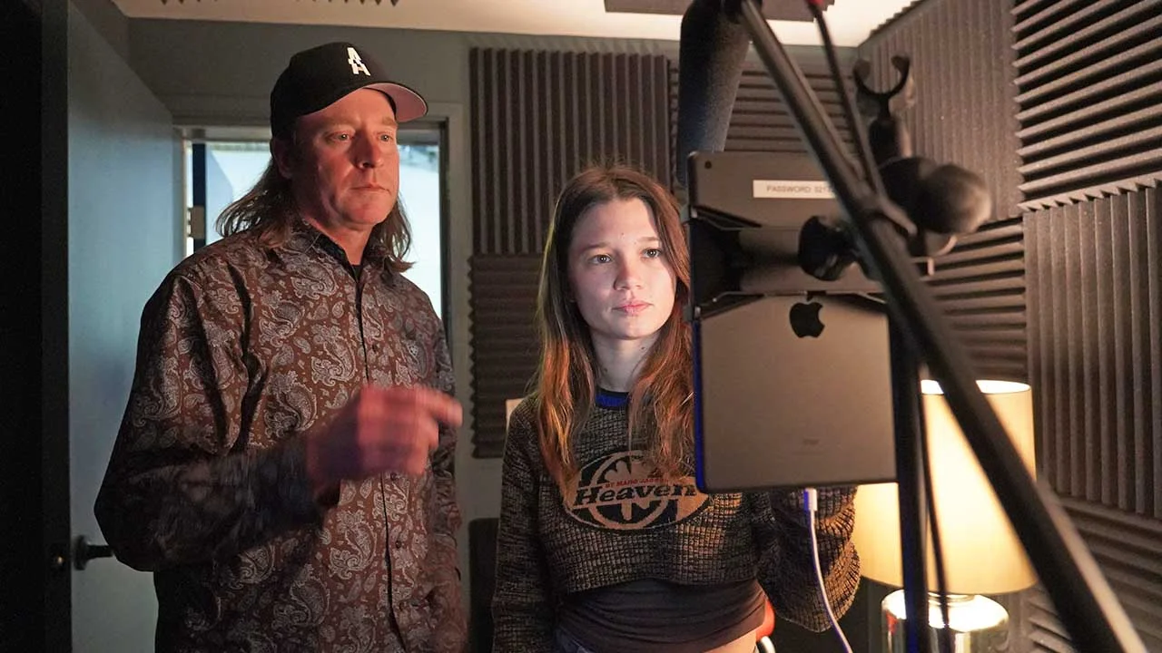 Two People in a Recording Studio Look at a Tablet Mounted on a Microphone Stand During a Session Surrounded by Acoustic Panels and a Lamp