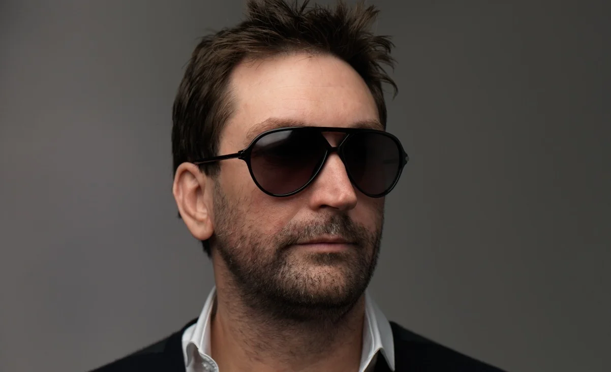 Portrait of a Man with Dark Sunglasses and Stubble Facing Slightly Right Against a Neutral Background Wearing a White Collared Shirt Beneath a Dark Sweater