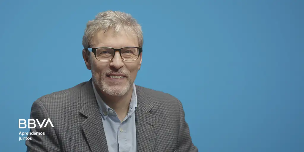 Portrait of a smiling man with gray hair and glasses in a checkered blazer against a blue background; BBVA logo and 'Aprendemos juntos' slogan in the corner.