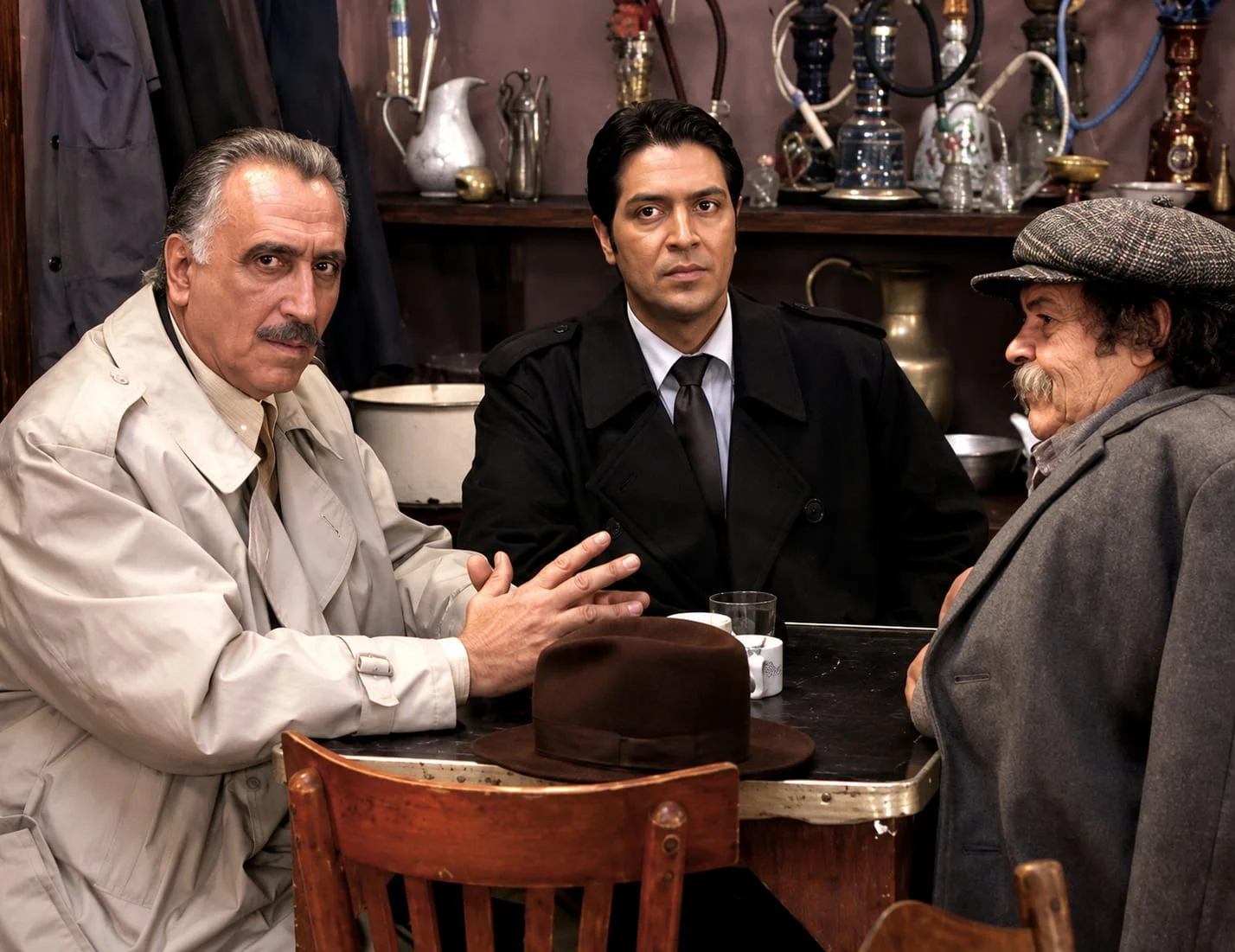 Three men sit around a wooden table in a dim, cluttered café or hookah bar, wearing coats; a brown fedora rests on the table in front of them.