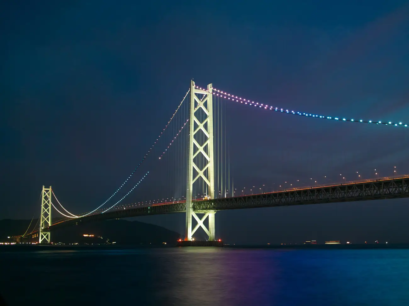 Akashi Kaikyo Bridge Night view of a suspension bridge lit with colorful LEDs, spanning calm water with reflections below.