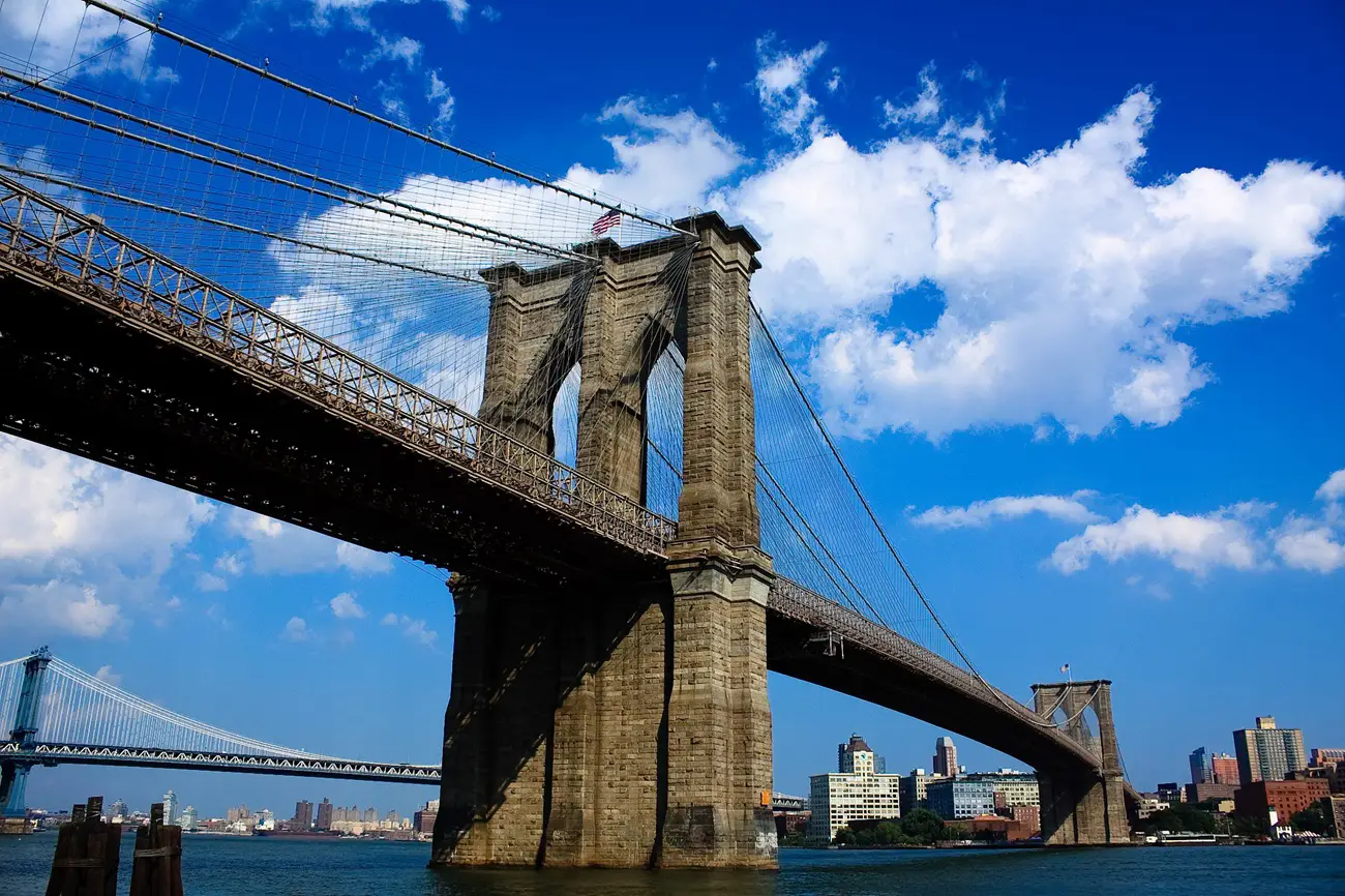 Brooklyn Bridge Brooklyn Bridge with stone towers, suspension cables, and a blue sky over the East River, with Manhattan skyline in the background