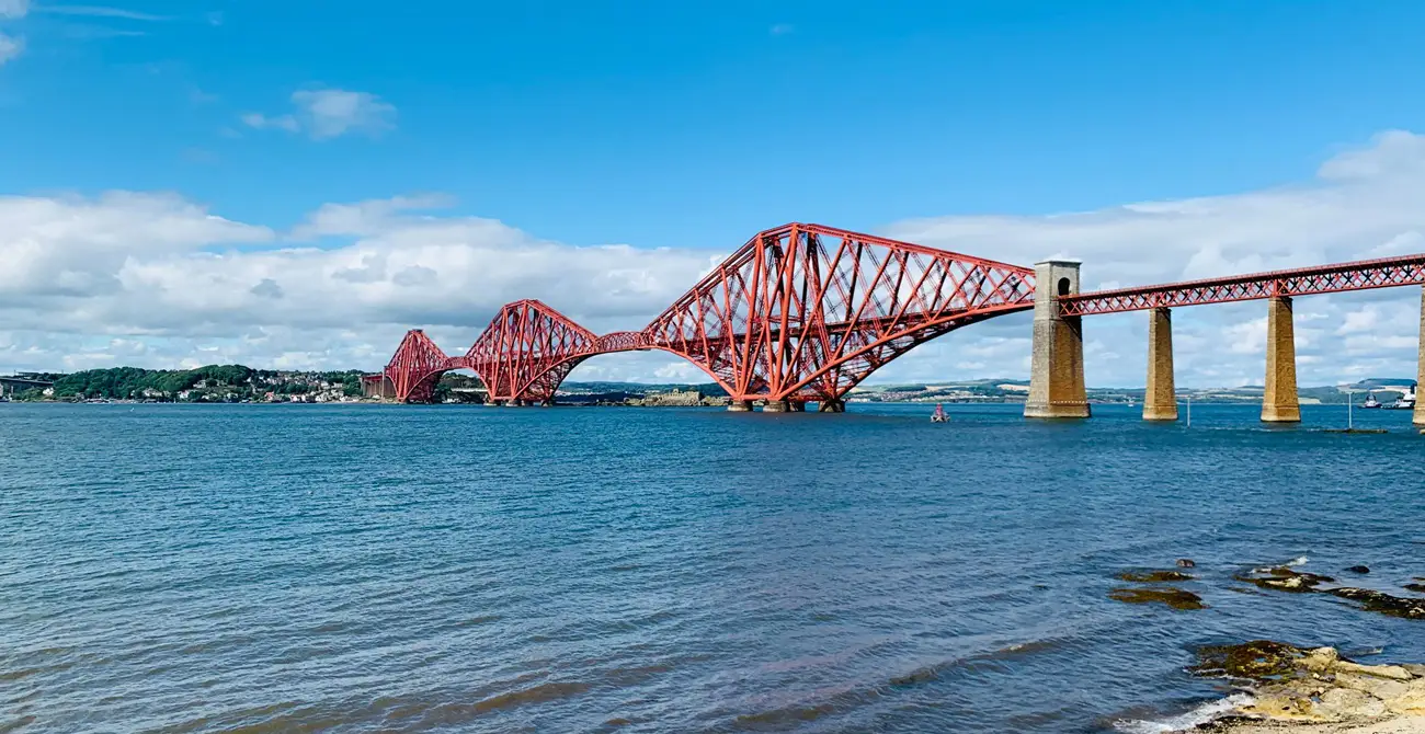 Forth Bridge Red cantilever railway bridge crossing blue water, with a distant shoreline and blue sky.