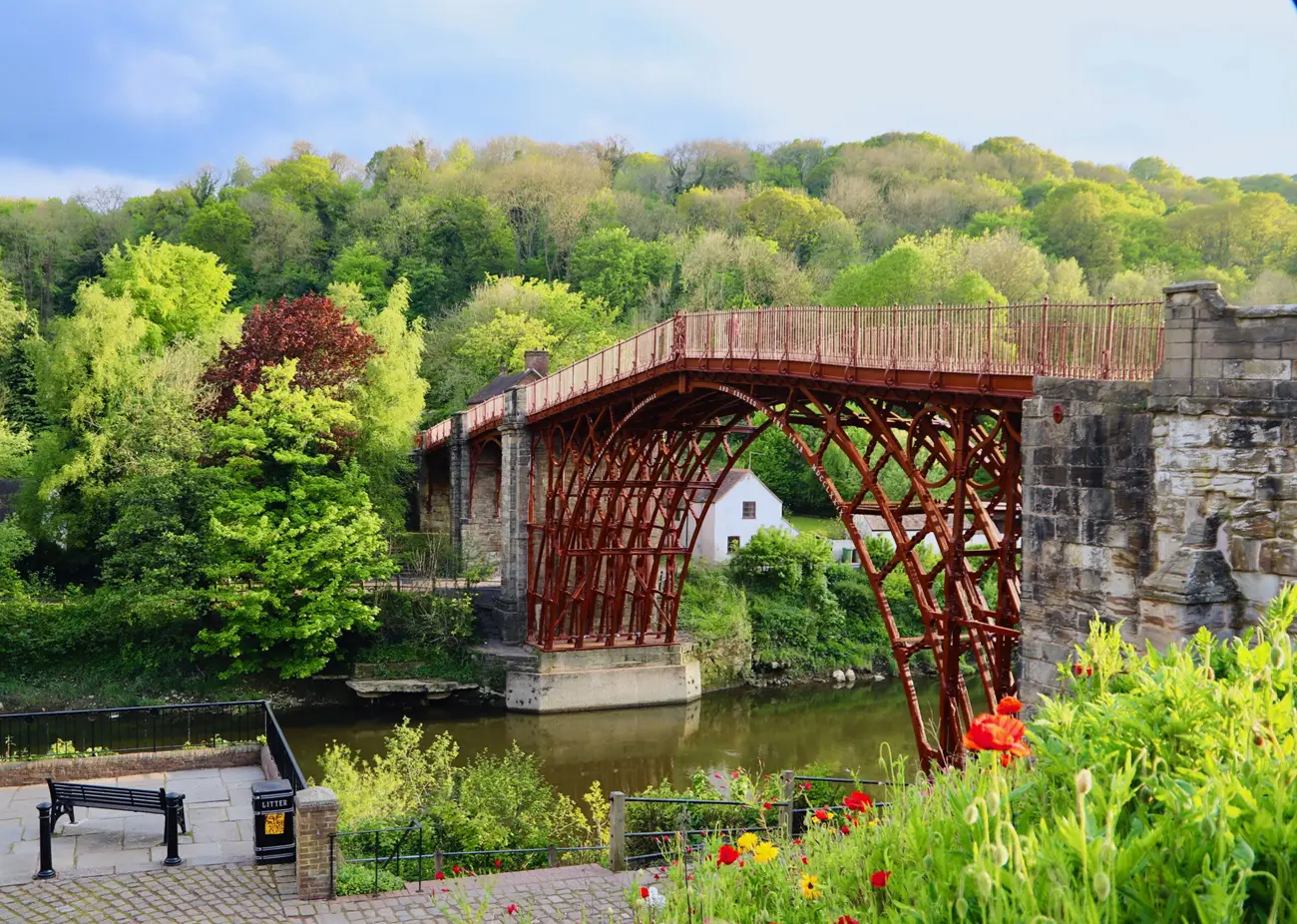 Iron Bridge Red wrought-iron lattice bridge crossing a calm river, with lush green trees and a small white house along the bank. foreground colorful flowers.