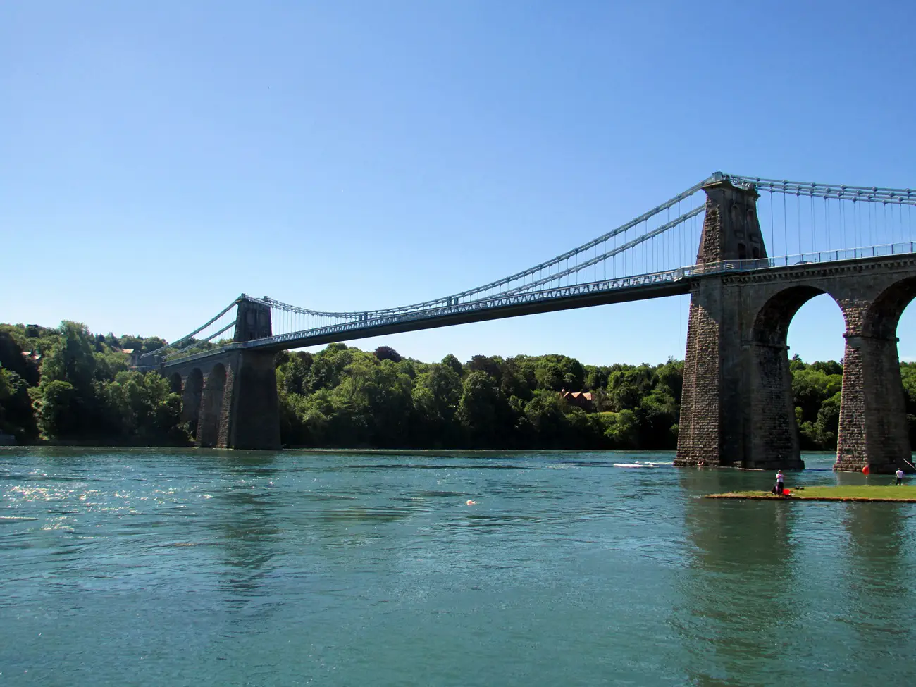 Menai Suspension Bridge Suspension bridge spanning a wide river with stone towers, flanked by trees on a sunny day.