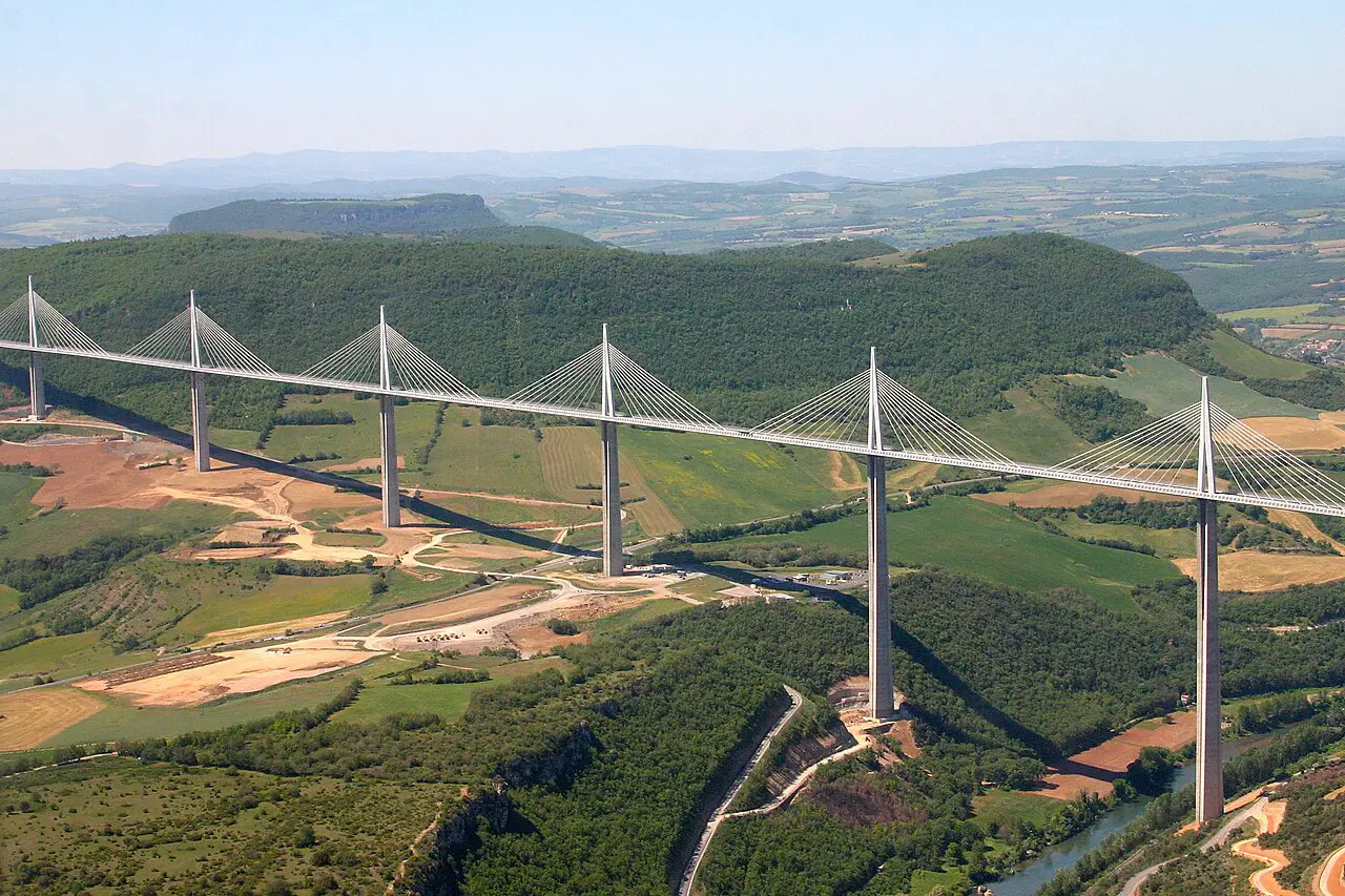 Millau Viaduct Cable-stayed bridge with tall white pylons spanning a green valley over a winding river. Reflective cables fan from each pylon to the deck amid rural fields and hills.