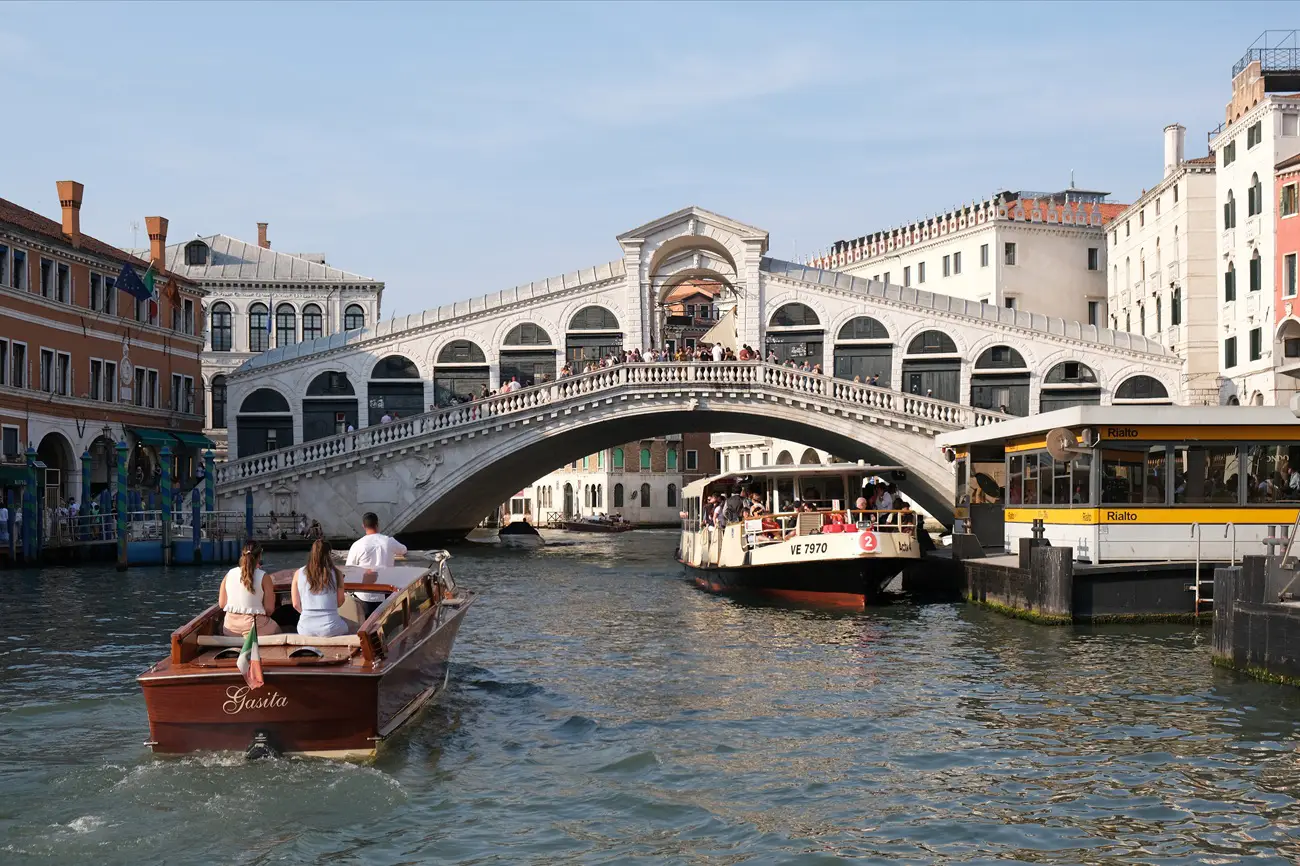 Rialto Bridge Wooden tour boat glides on a Venetian canal under the arches of the Rialto Bridge, with people aboard and city buildings beside.