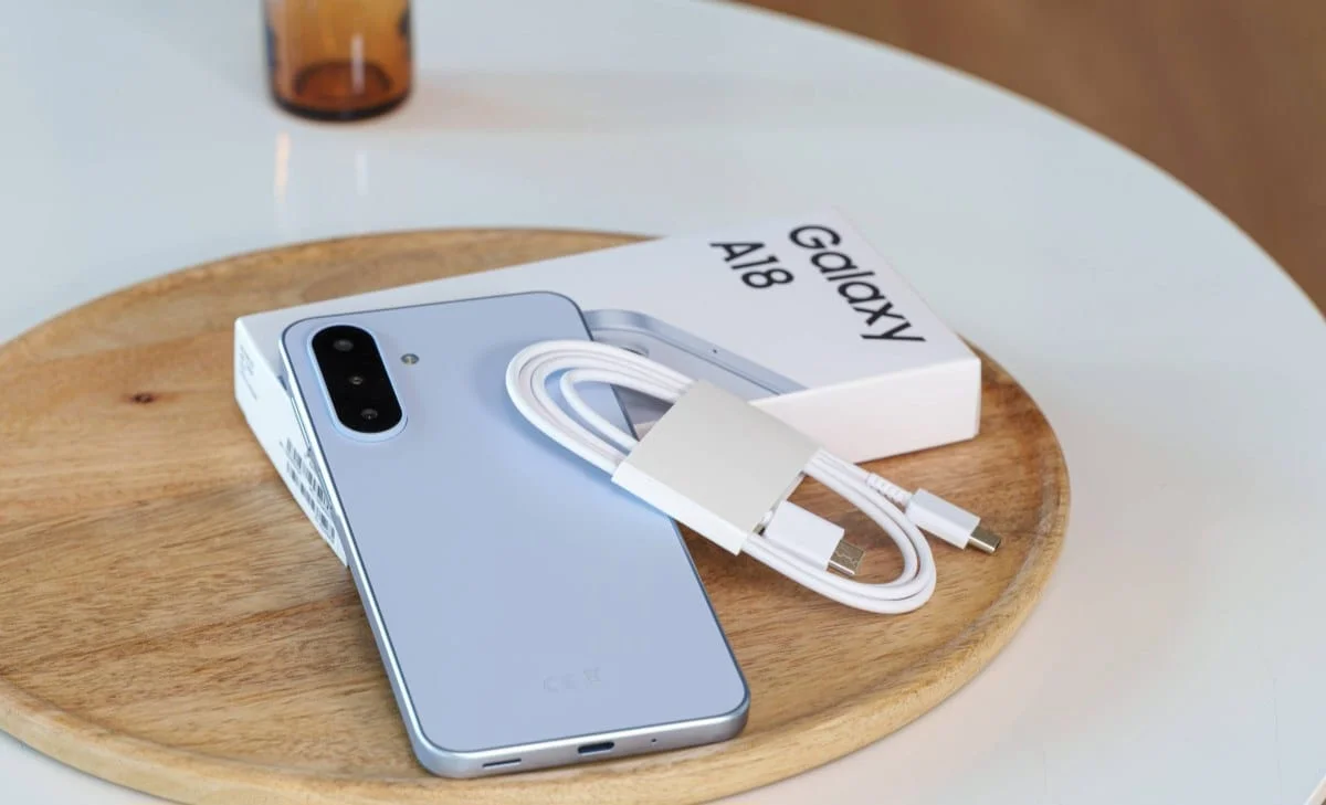 gsmarena_016 Blue Samsung Galaxy A18 smartphone with a white charging cable and adapter resting on its box on a round wooden tray.