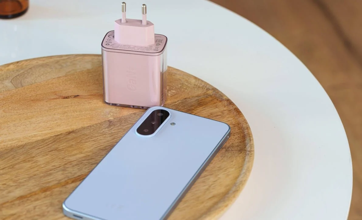 gsmarena_017 Light blue smartphone with a large camera module rests on a wooden cutting board beside a pink plug-in power adapter.