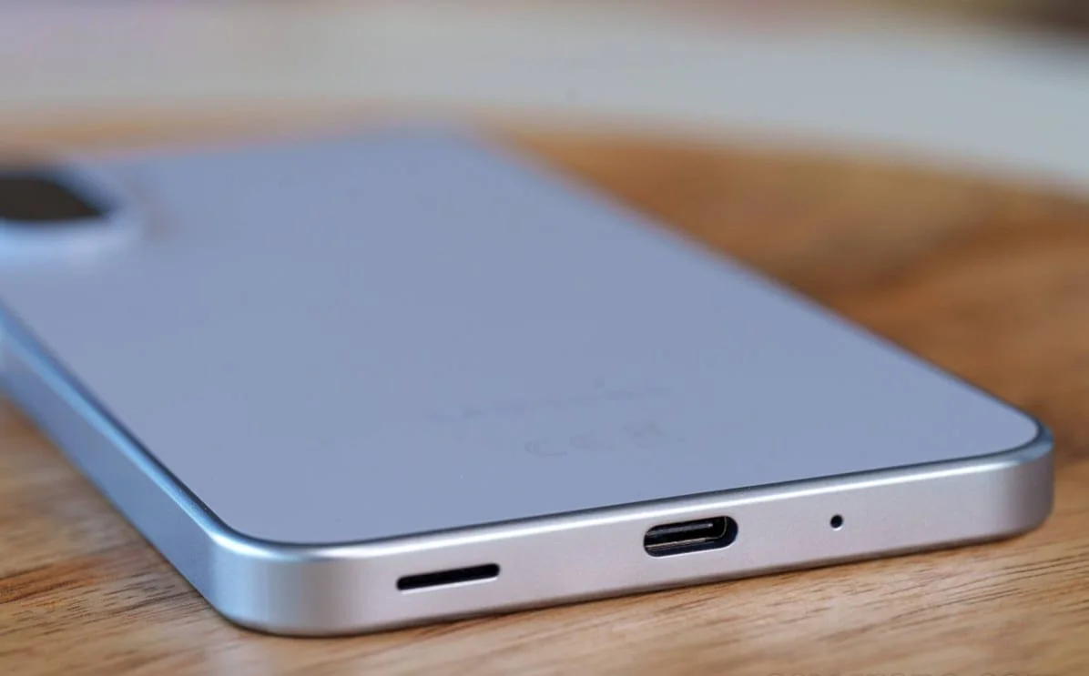 gsmarena_025 Close-up of a silver smartphone lying on a wooden surface, showing the bottom edge with a USB-C port and speaker grille.