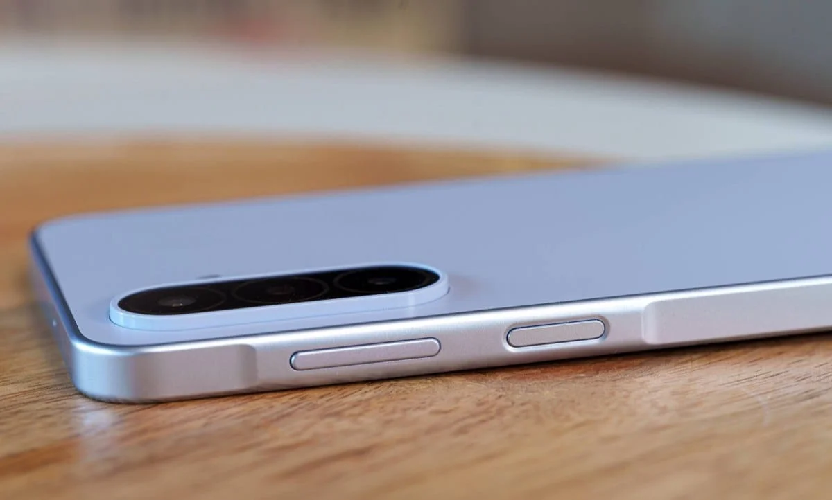 gsmarena_027 Close-up of a silver smartphone on a wooden surface, highlighting its triple-camera rear module and side buttons