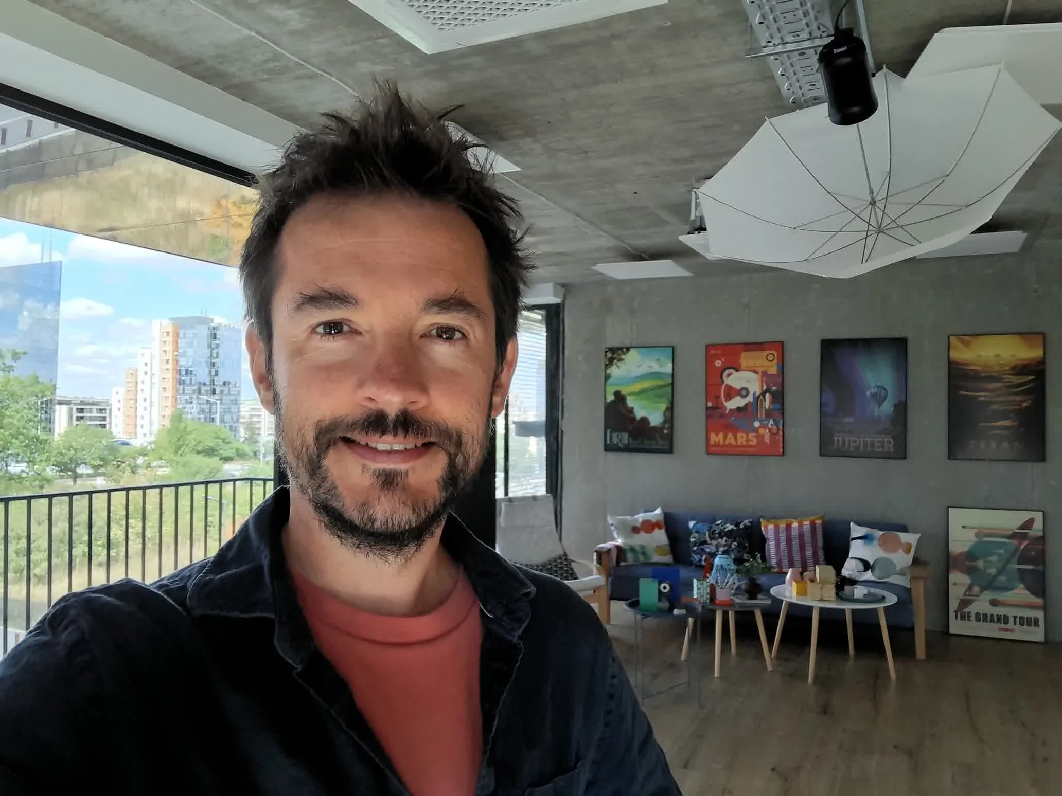 gsmarena_3001 Selfie of a smiling man with short dark hair and stubble indoors, with colorful posters on the wall behind him and a window on the left.