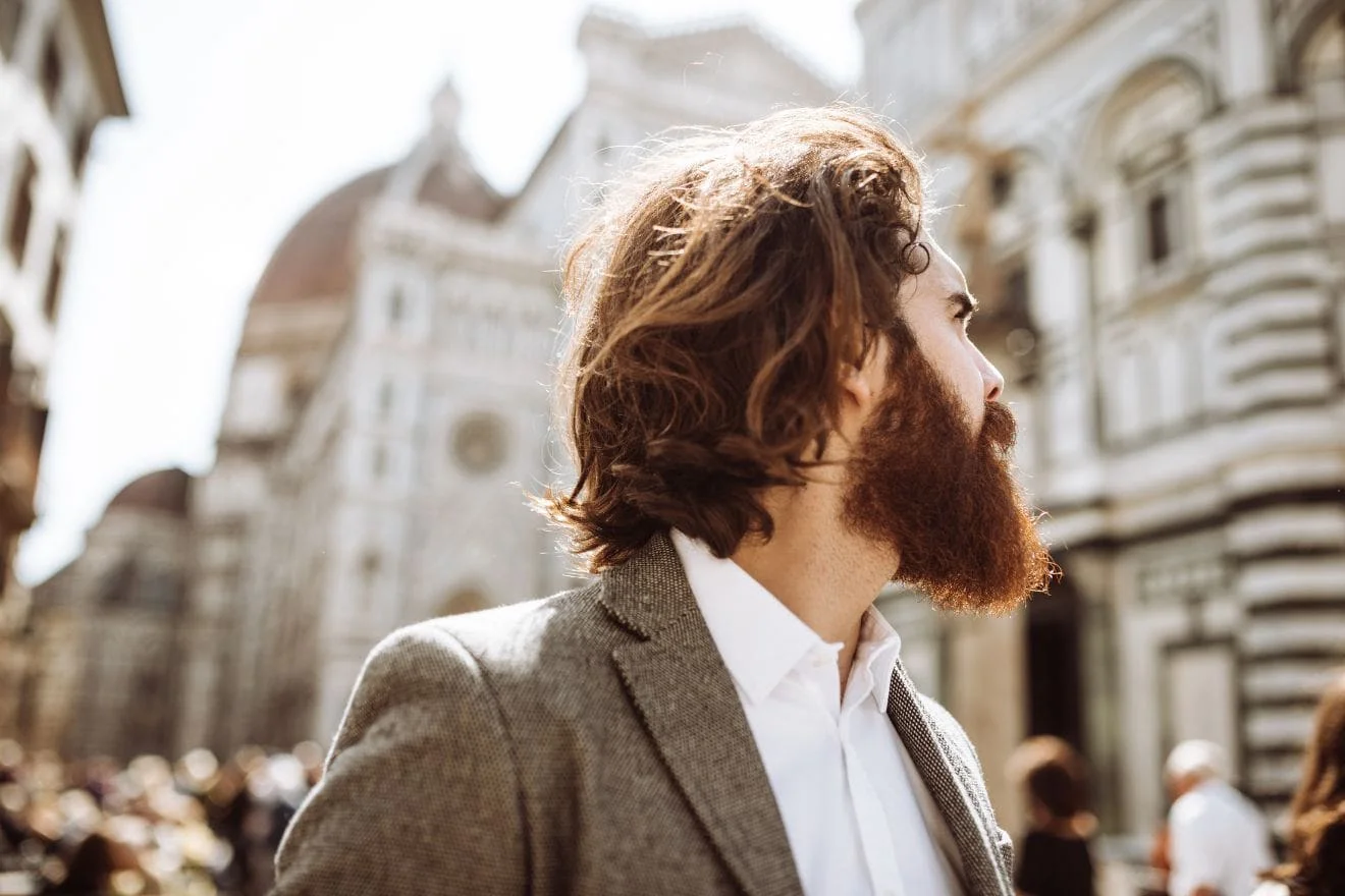 longhair Side profile of a bearded man with wavy brown hair wearing a gray blazer on a sunny city street with historic buildings in the background.