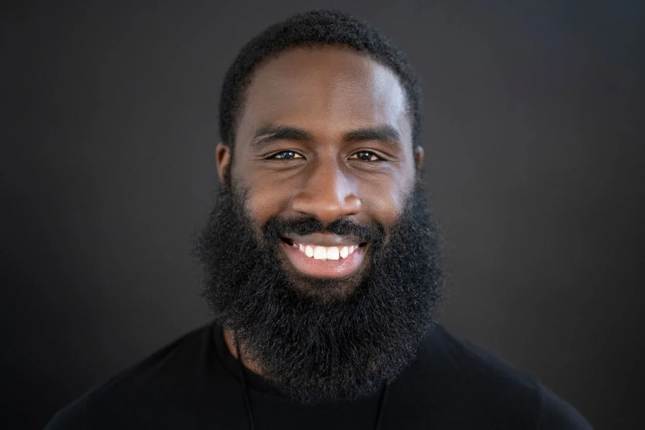thickbeard Close-up portrait of a smiling man with a thick beard and short hair against a dark background.