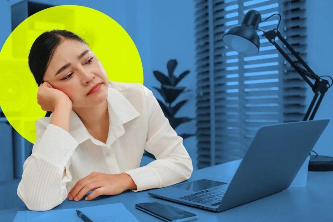 work-from-home-grim-effect-brain Woman resting her head on her hand at a desk with a laptop, looking fatigued or bored in an office setting.